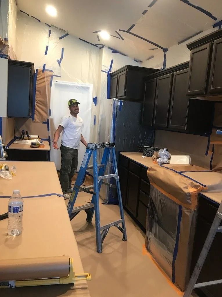 A person in a white shirt and cap works in a kitchen with newly painted dark cabinets and surfaces protected by paper.