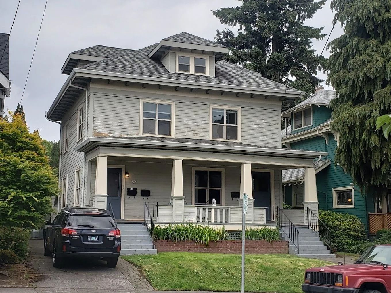 A light-gray, two-story foursquare house with a covered front porch, a shingled roof, and a black SUV parked in the driveway.