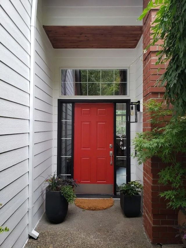 A front entrance with a bright red door, black trim, a transom window, wood ceiling detail, and potted plants on either side.