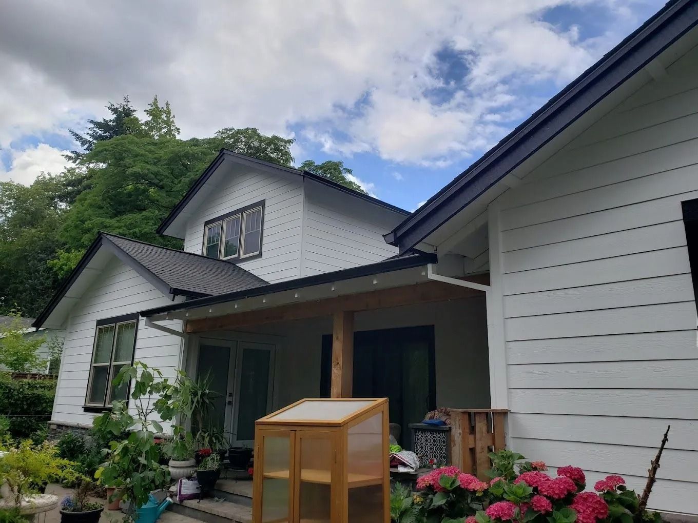 A white farmhouse with black trim, a covered porch, a small wooden greenhouse, and pink flowers against a cloudy sky.
