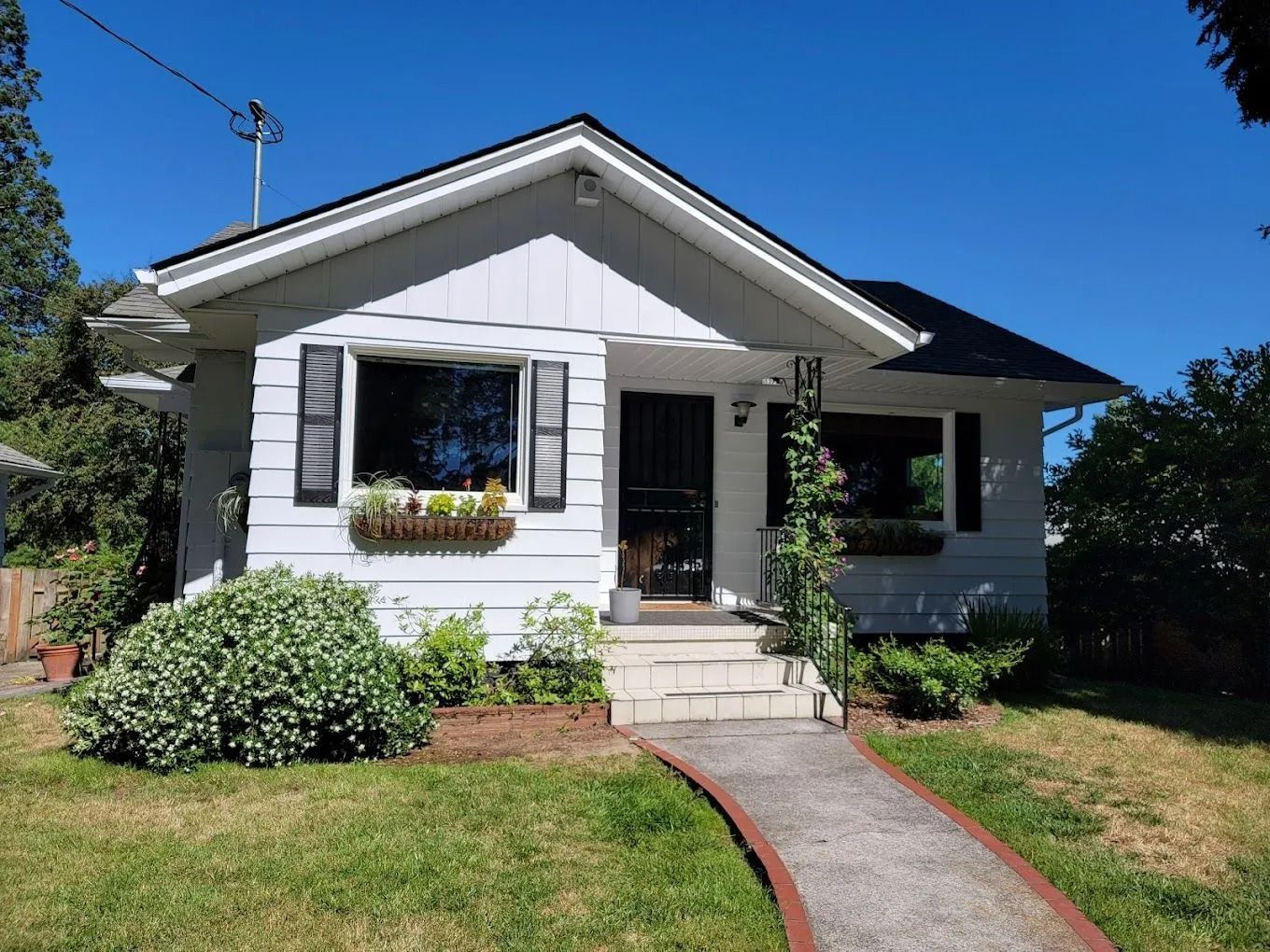 A single-story white house with dark shutters, a front porch, and a walkway, set against a clear blue sky.