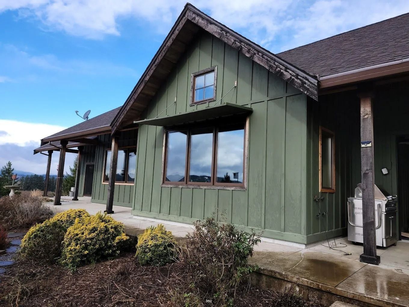 A green, board-and-batten single-story house with a wooden porch, dark roof, and large front windows under a cloudy sky.