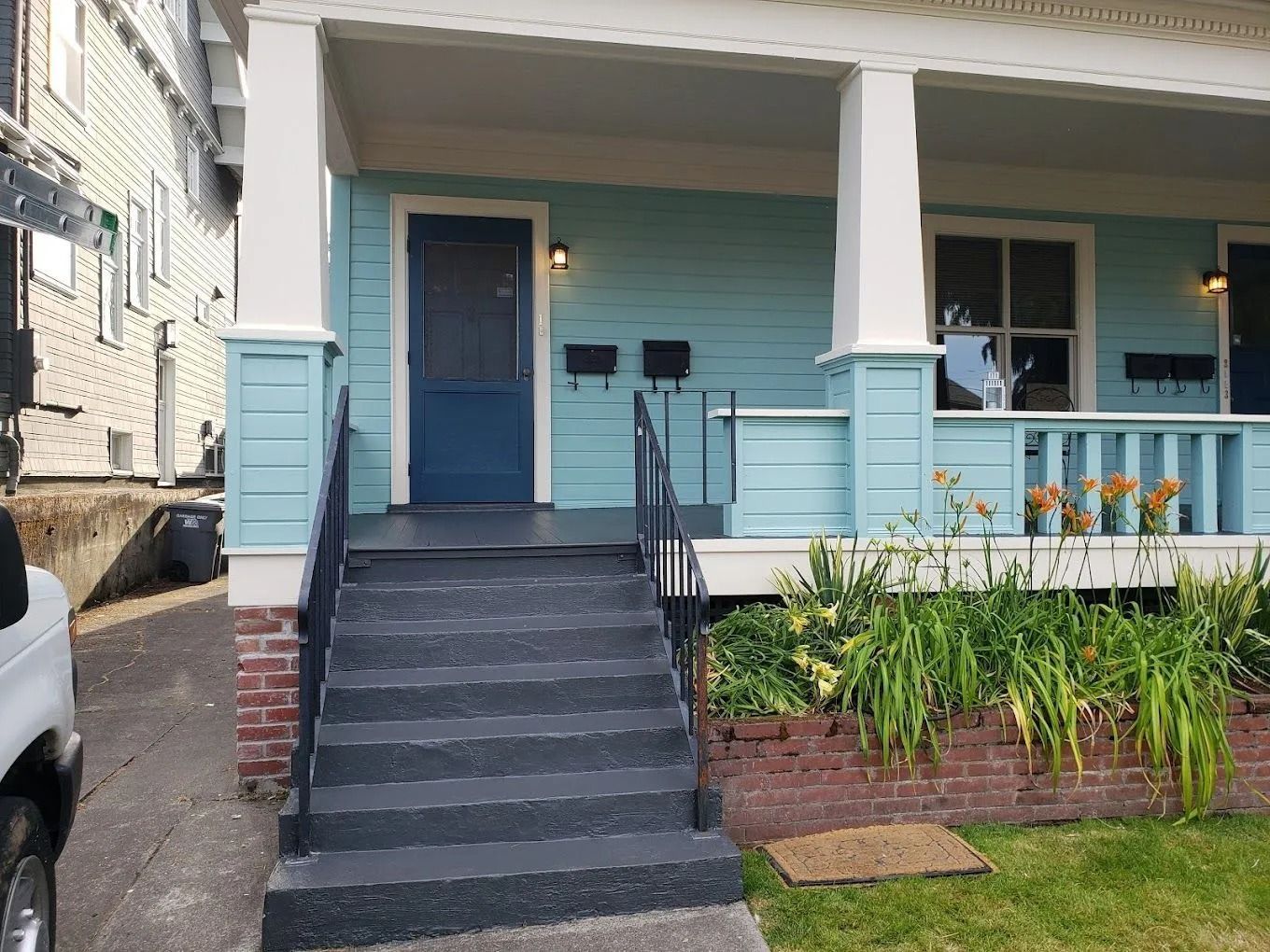 A light blue house front porch with a blue door, black metal railings, and a small brick planter with green plants.