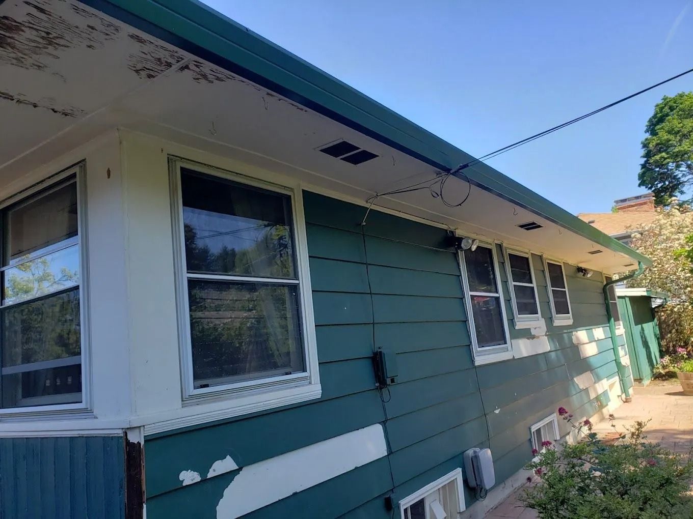 A side view of a dark teal house with peeling paint, several windows, and a white soffit under a clear blue sky.