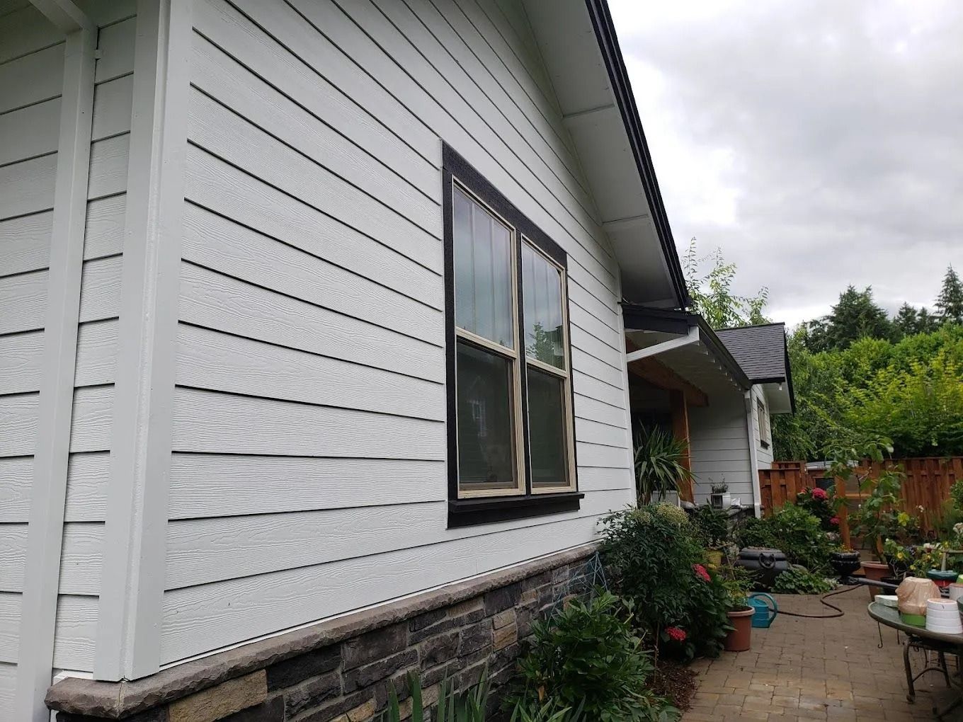 A side view of a white house with horizontal siding, dark window trim, and a stone facade foundation beside a patio.
