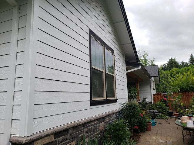 A side view of a white, lap-sided house exterior with a black-framed window and stone veneer base, beside a paved patio.