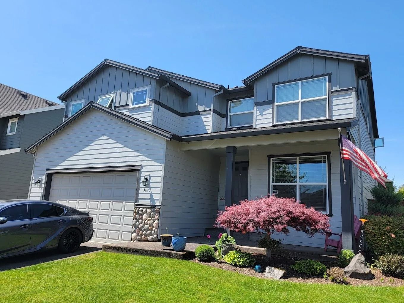 A two-story grey suburban house with a two-car garage, a small landscaped front yard, and an American flag on the porch.