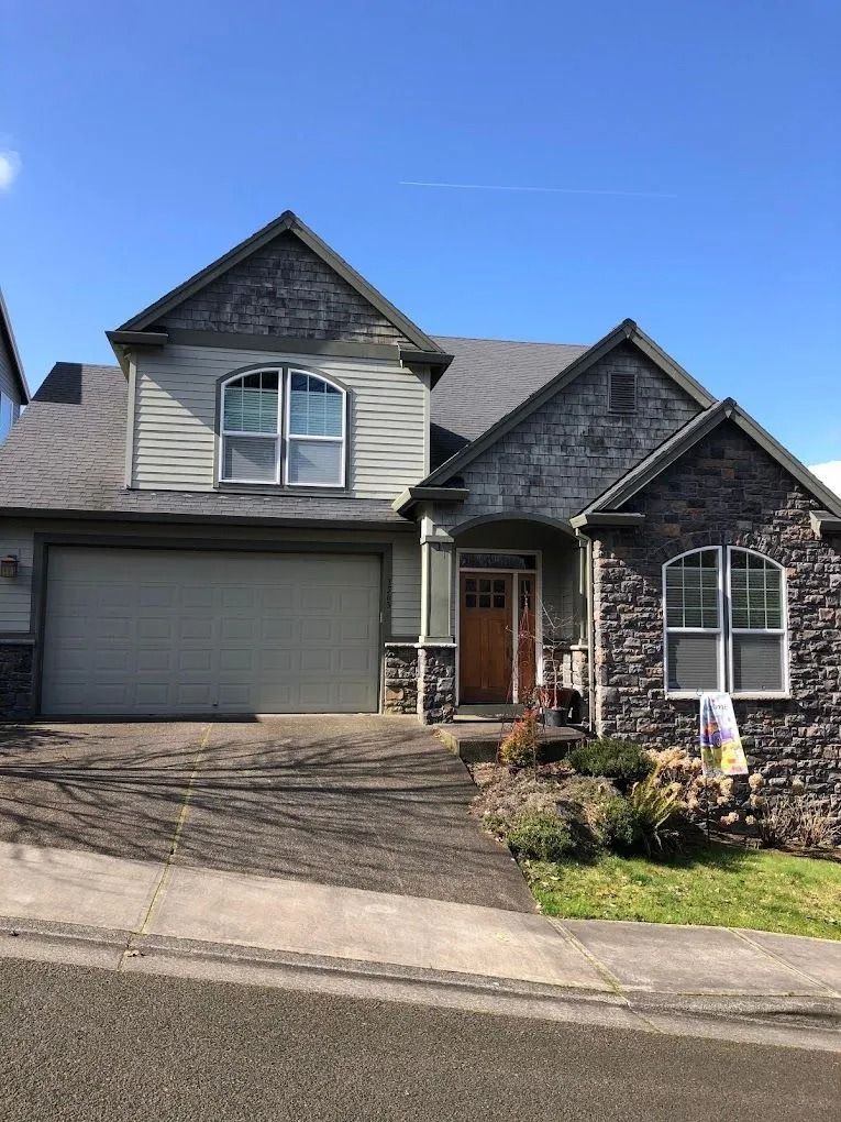 A two-story suburban house with a gray stone and beige siding facade, a front-facing garage, and a wooden front door.
