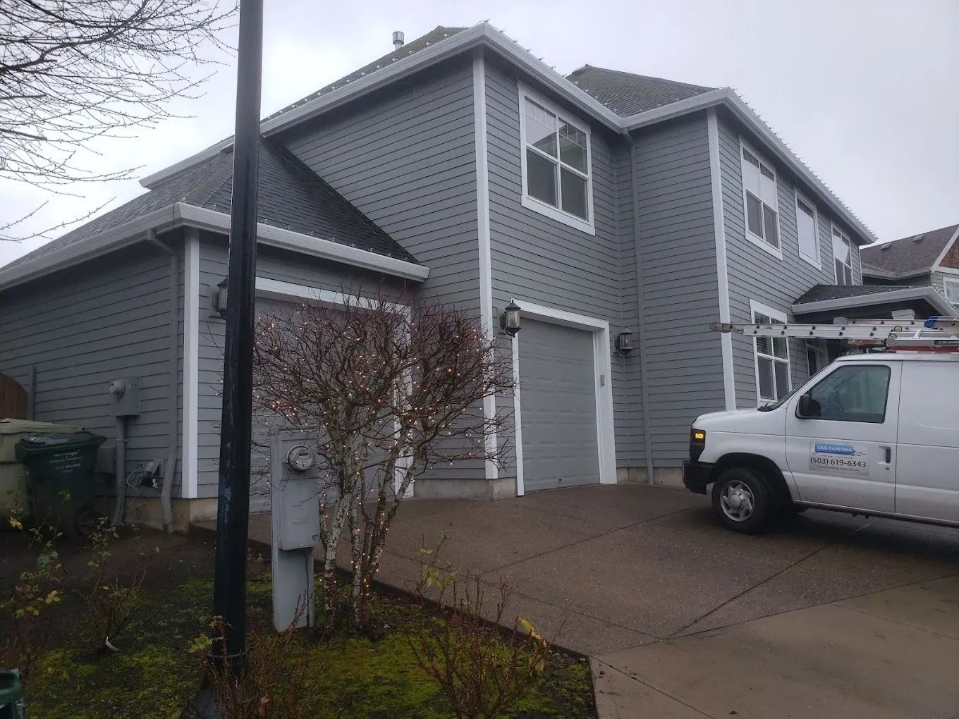 A gray two-story house with two garage bays and a service van parked in the driveway on an overcast day.