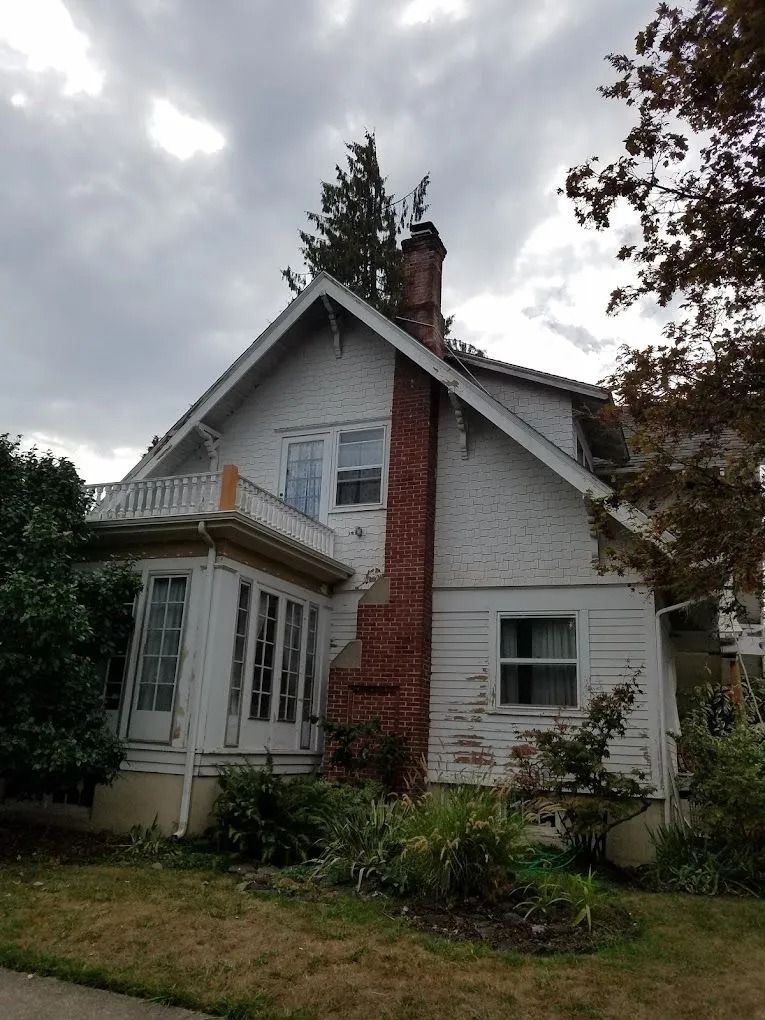 A two-story house with white siding, a red brick exterior chimney, a sunroom with large windows, and a railed porch.