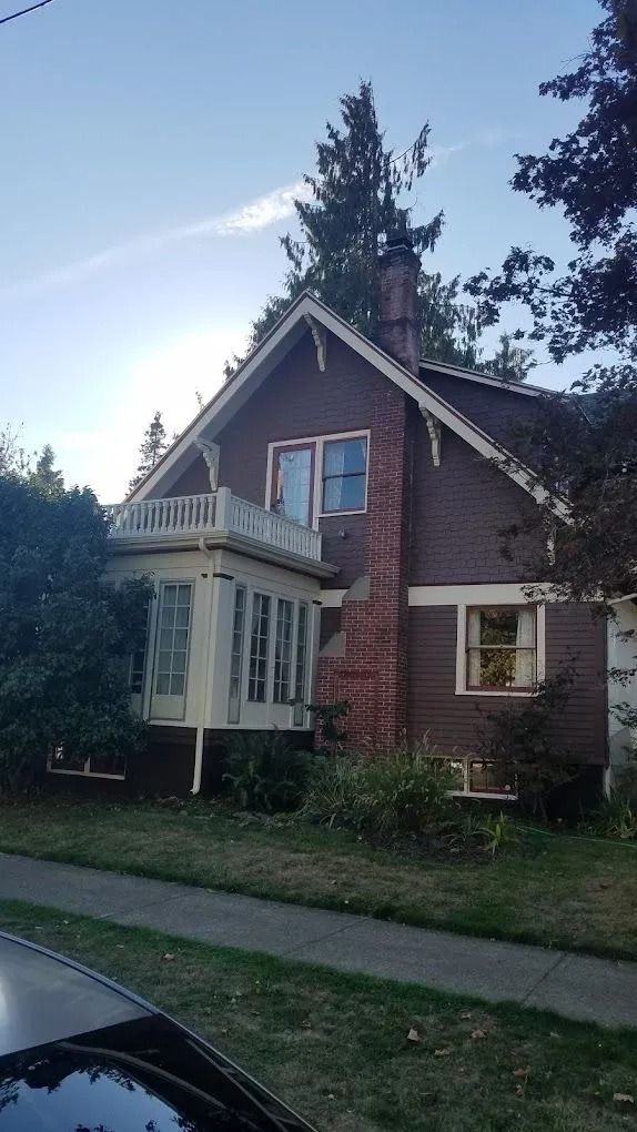 A two-story, dark brown house with a white sunroom, a brick chimney, and a small balcony under a gabled roof.