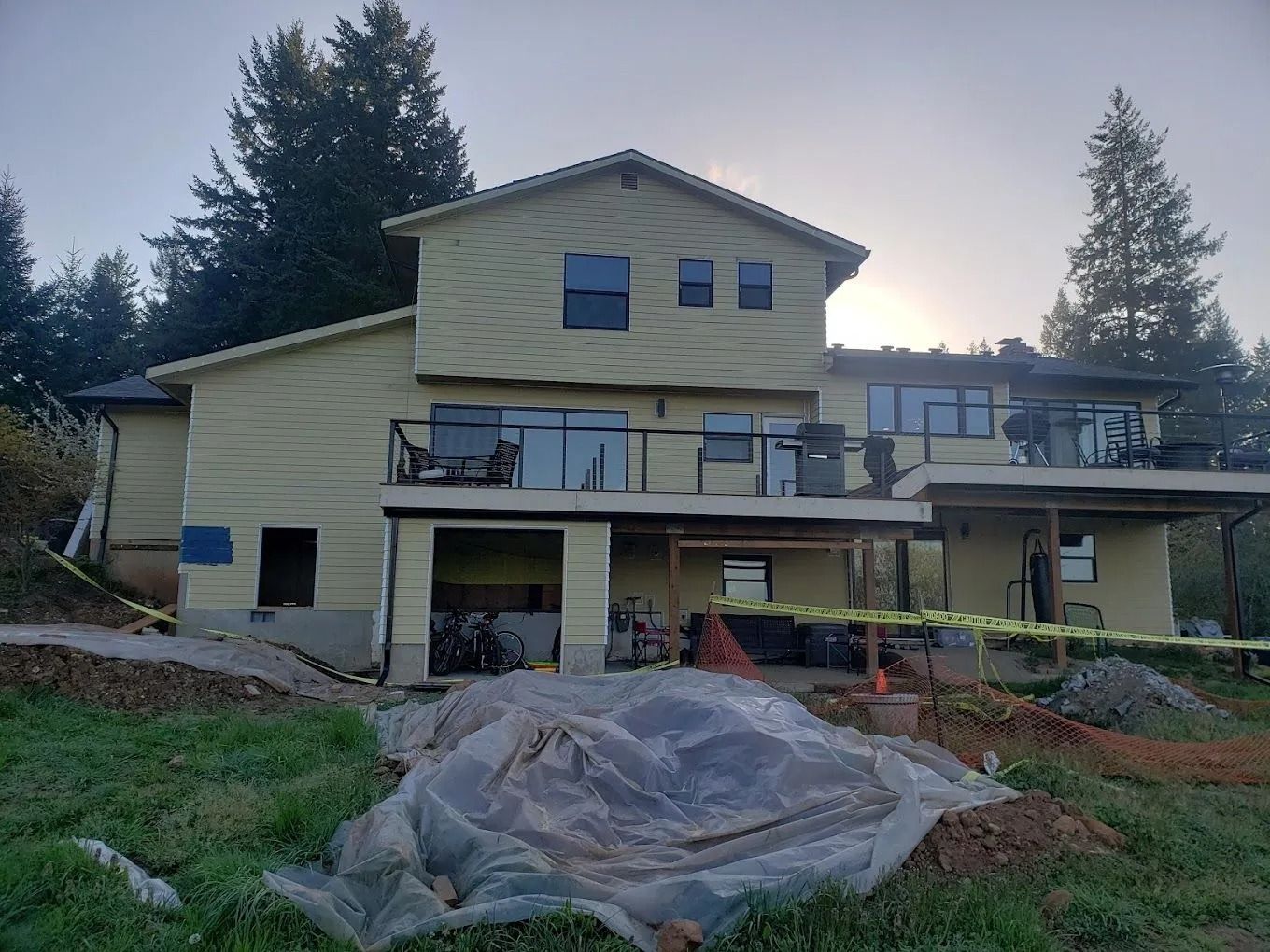 A multi-story house with yellow siding and a deck under construction, viewed from a yard with plastic ground sheeting.
