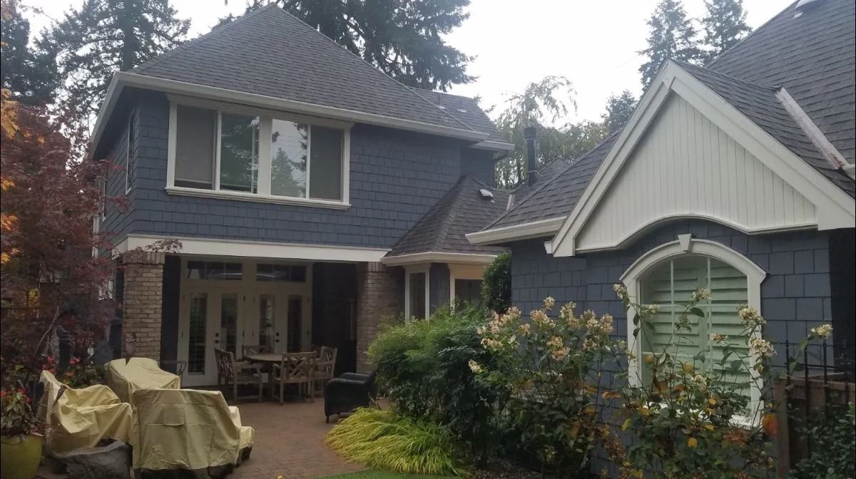 A two-story blue house with dark shingles and white trim, featuring a patio area with furniture and surrounding greenery.