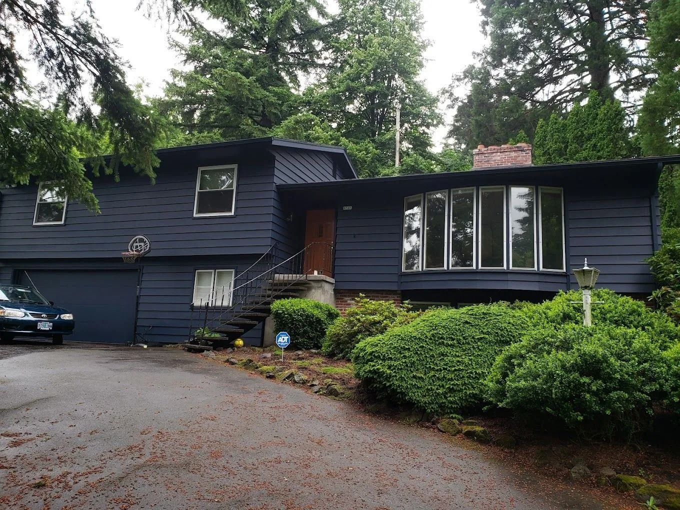 A split-level house painted dark blue with a large bay window, surrounded by trees and overgrown bushes.