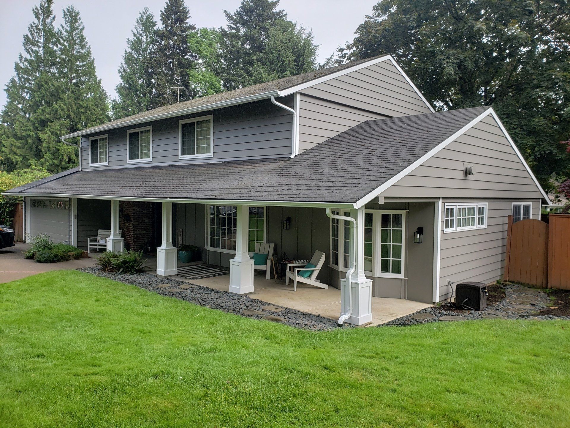 A two-story grey house with a large covered patio, white support pillars, and a green lawn in the foreground.