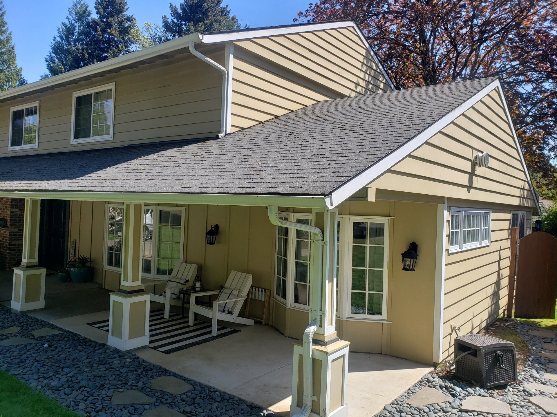 A tan two-story house with a covered patio, featuring white pillars, grey shingles, and outdoor lounge chairs.