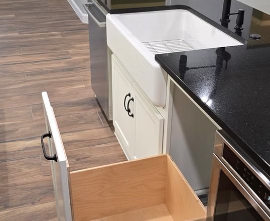 Kitchen island with open drawer, farmhouse sink, black countertop, light cabinets, and wood-look flooring.