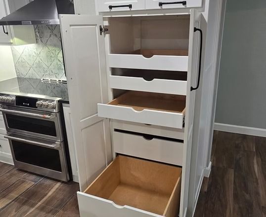 White kitchen cabinet with pull-out drawers, open. Located next to a stainless steel oven.