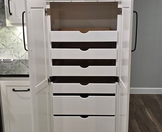 White kitchen pantry cabinet with pull-out drawers, open. Black handles on doors.