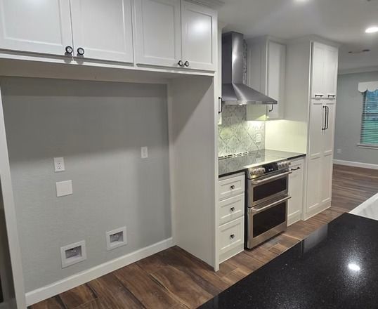 White kitchen cabinetry and appliances, wood flooring, grey wall, built-in oven, range hood.