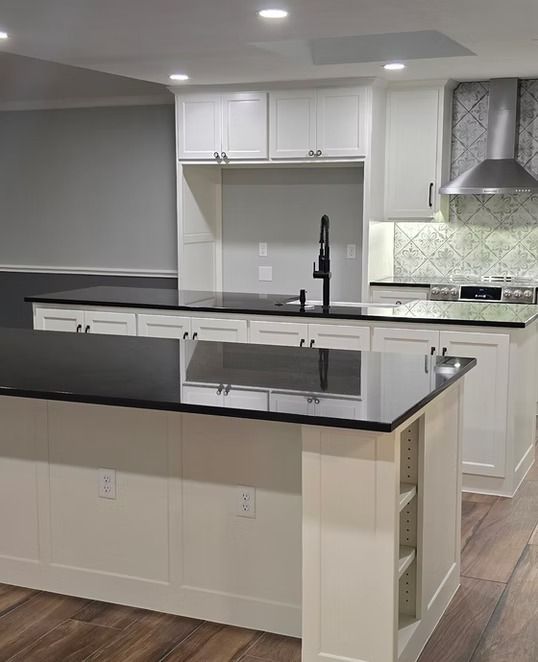 Modern white kitchen with black countertops and black faucet, includes island with shelving, white cabinetry, wood floor.