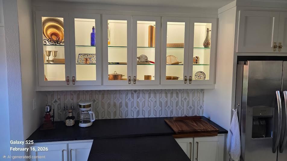 White upper cabinets with glass doors and internal lighting above a black countertop and patterned backsplash.
