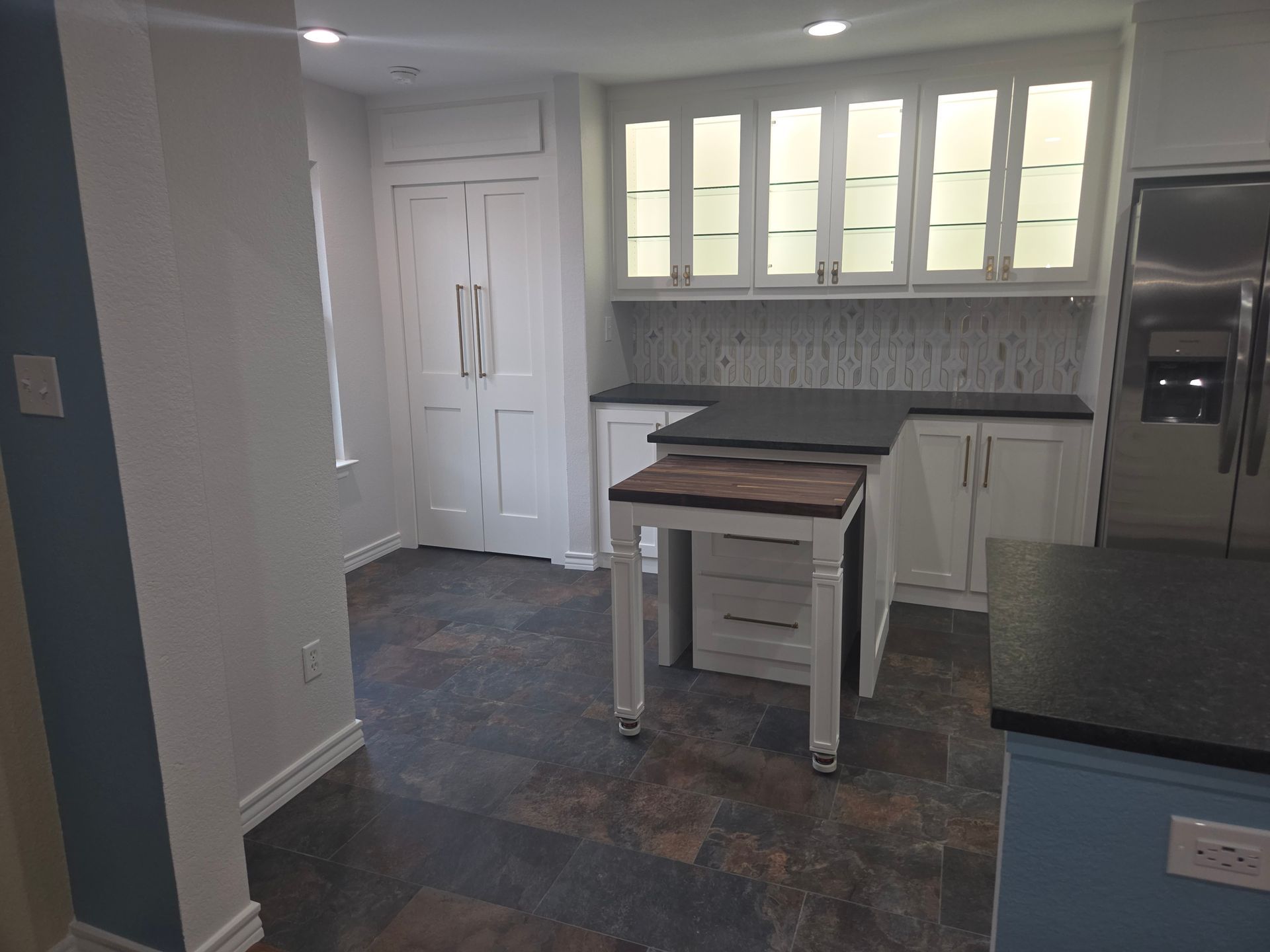 A kitchen with white cabinets, glass-front upper cabinets, a rolling butcher block island, and dark countertops.