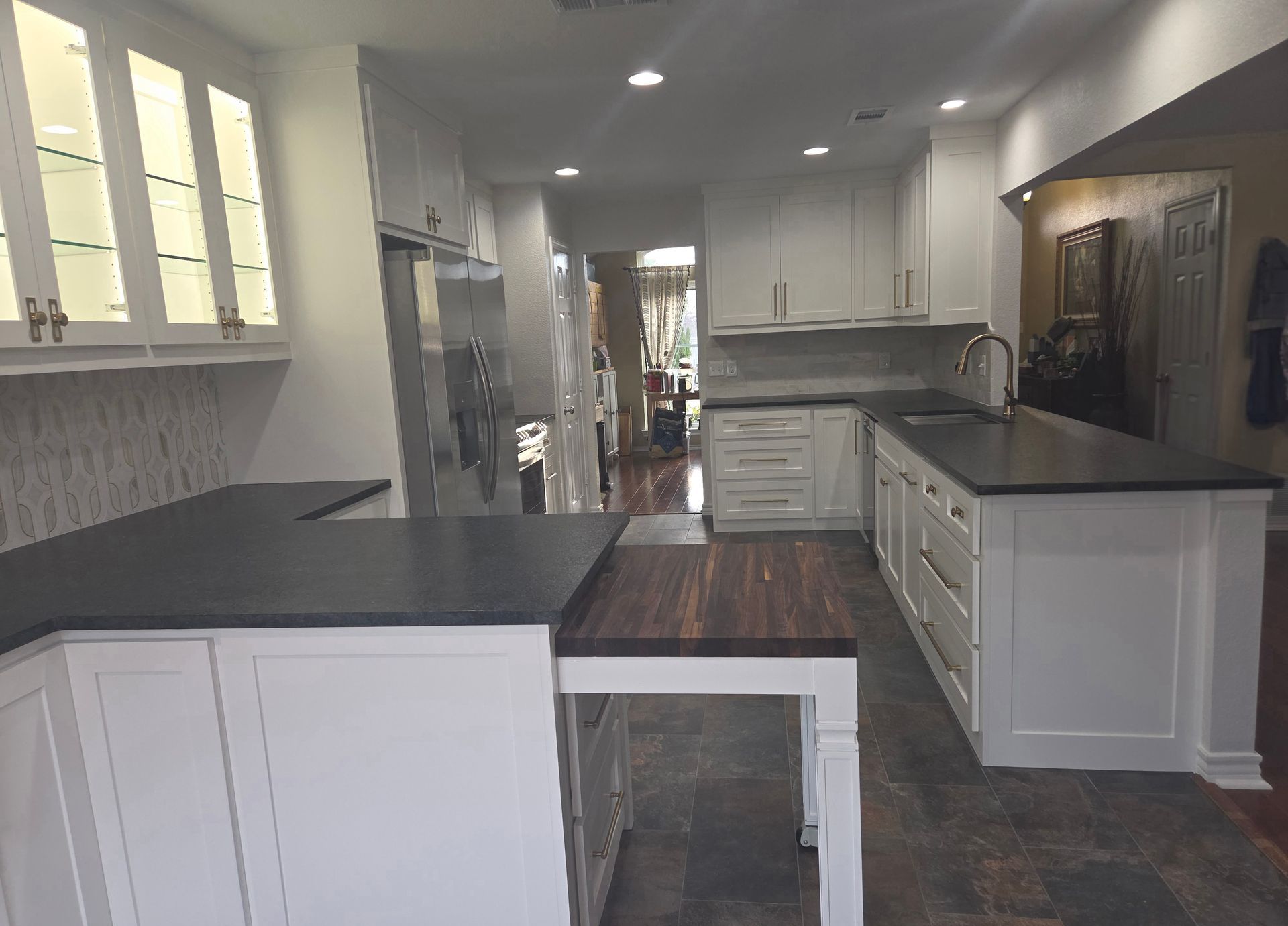 A white kitchen featuring dark countertops, a wooden prep table extension, glass-front cabinets, and tile flooring.