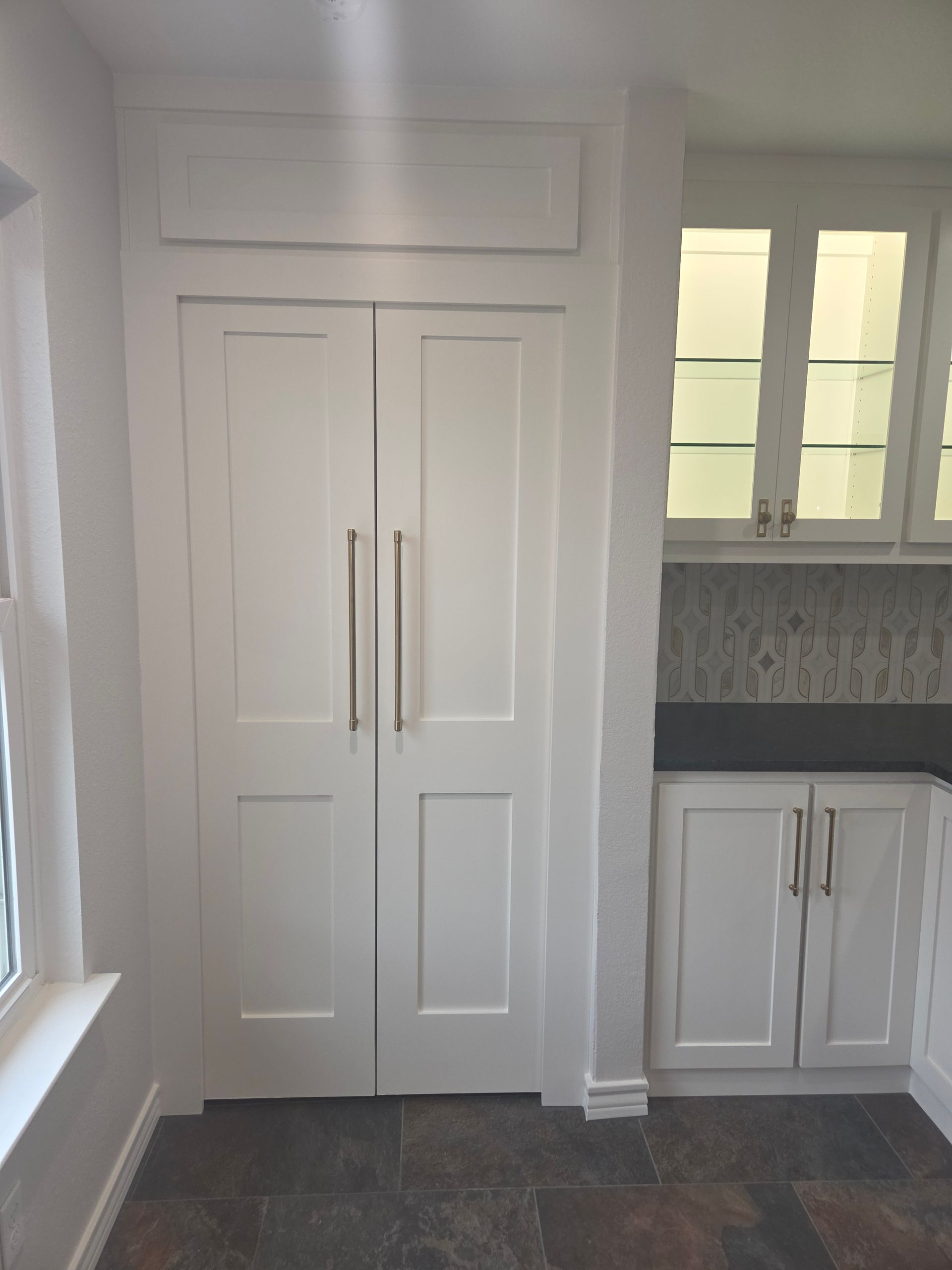 Tall white paneled cabinet doors with long gold handles next to a kitchen counter with glass-front upper cabinets.