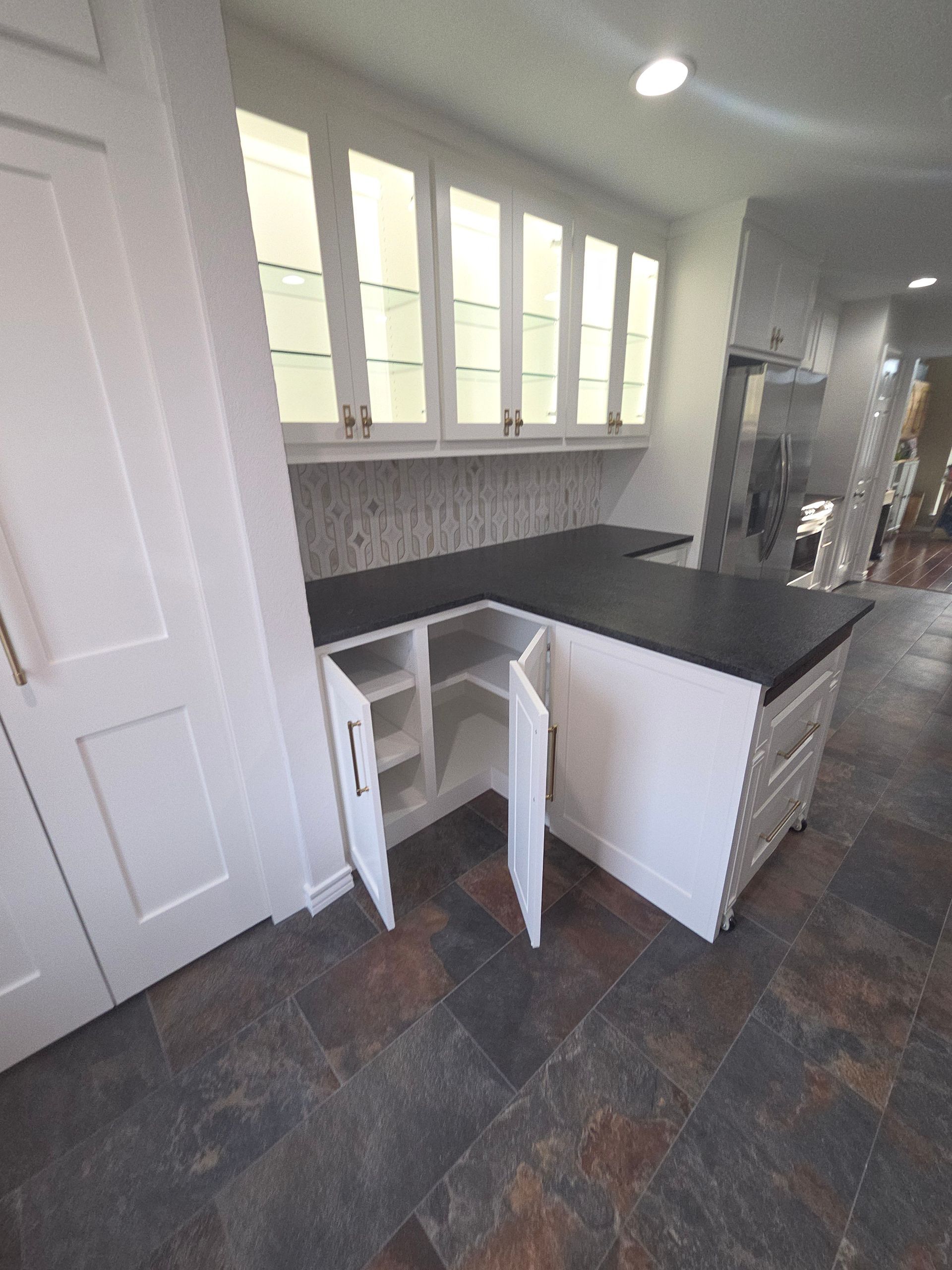 A white kitchen cabinet unit with a black countertop and glass-front upper cabinets, with corner base doors standing open.