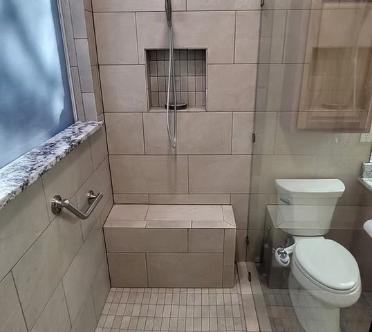 Bathroom shower stall with bench and grab bar, featuring neutral-toned tiles, glass door, and toilet.