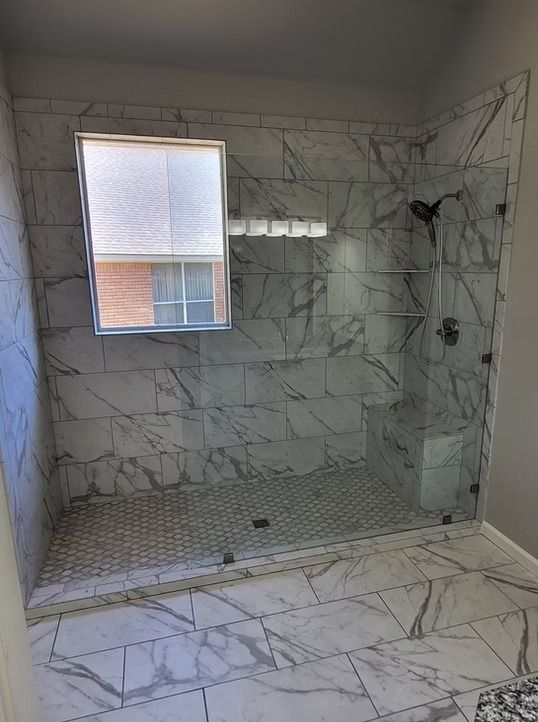 Spacious shower with marble-like tile, glass doors, and a window. Black shower fixtures and a gray floor.
