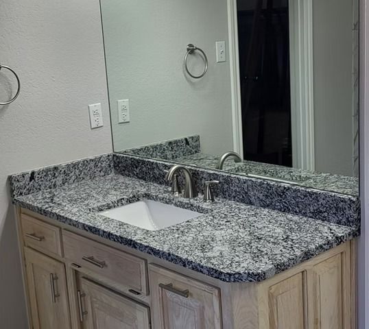 Bathroom with granite countertop, sink, mirror, and light-colored wooden cabinets.