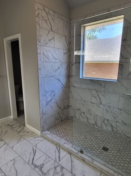 Bathroom with white marble tile shower and floor, clear glass enclosure, and window.