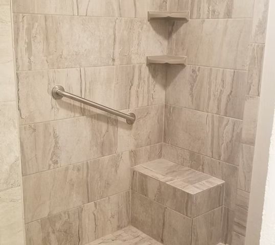 Tiled shower with grab bar, built-in seat, and corner shelves. Neutral tones and stone-like texture.