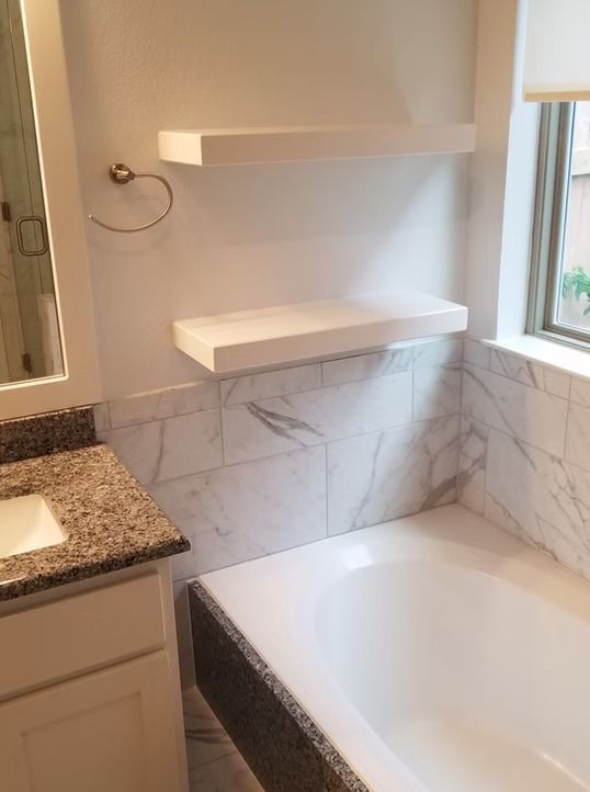 Bathroom with white bathtub, marble tile, floating shelves, and window.