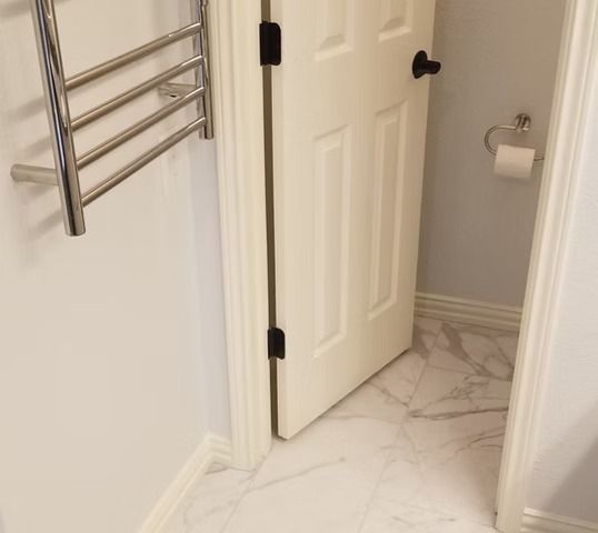 White bathroom door open, revealing toilet and marble floor. Chrome towel rack on left.