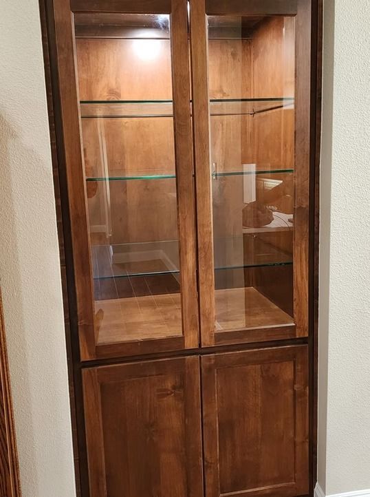 Wooden cabinet with glass-doored upper section and solid-doored lower section. Shelves and items inside.