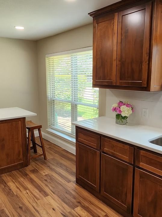Kitchen with brown cabinets, white countertops, window with blinds, and wooden floor.