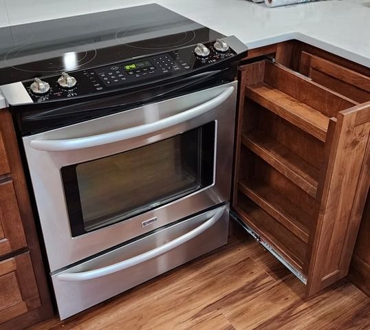 Stainless steel oven with pull-out spice rack in a kitchen with wooden cabinets.