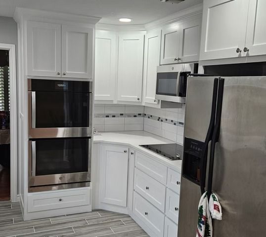 White kitchen cabinets with built-in ovens, microwave, and stainless steel refrigerator on gray tiled floor.
