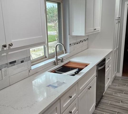 White kitchen with quartz countertop, stainless steel sink and faucet, white cabinets, and a window.