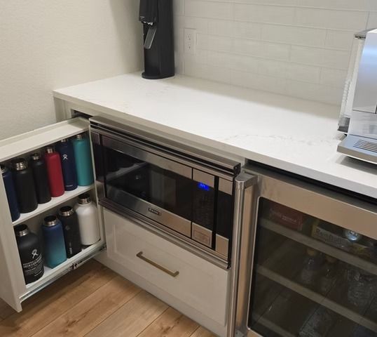 Built-in kitchen area with microwave, small fridge, and water bottles in a cabinet. White countertop, wood floor.
