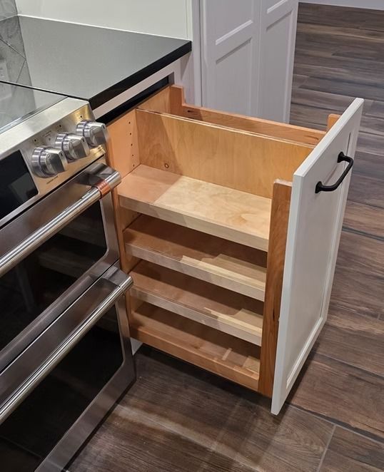 Cabinet drawer with spice rack pulled out next to a kitchen oven. White cabinet, wooden shelves, dark handle.