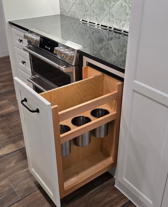 White cabinet with pull-out utensil holder beside a stainless steel oven and stovetop in a kitchen.