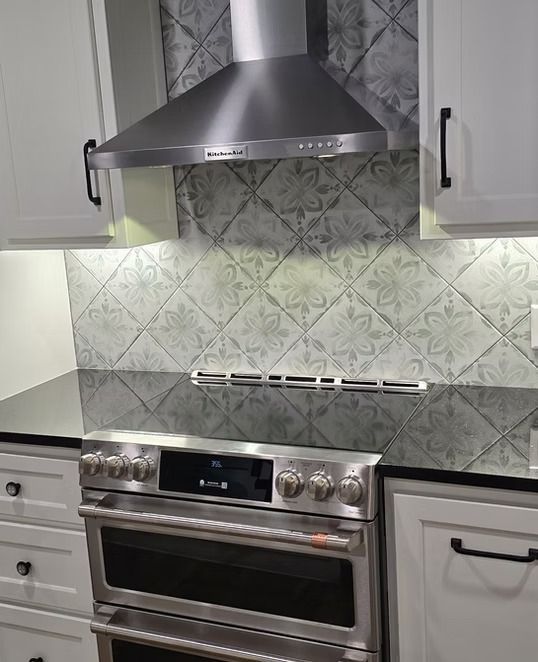 Stainless steel range, range hood, and floral tile backsplash in kitchen. White cabinets, black countertop.