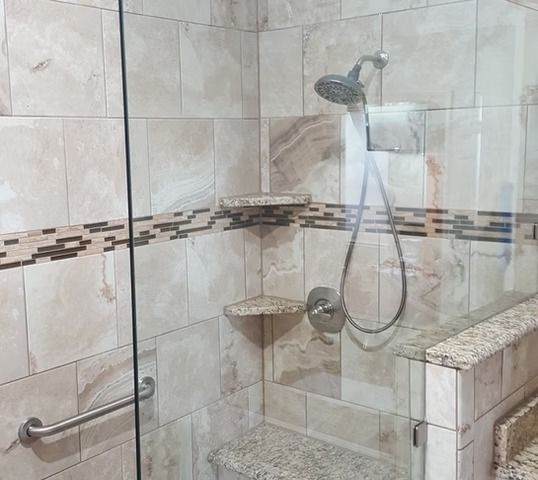 Shower stall with beige tiles, glass door, and handrail; built-in shelves.