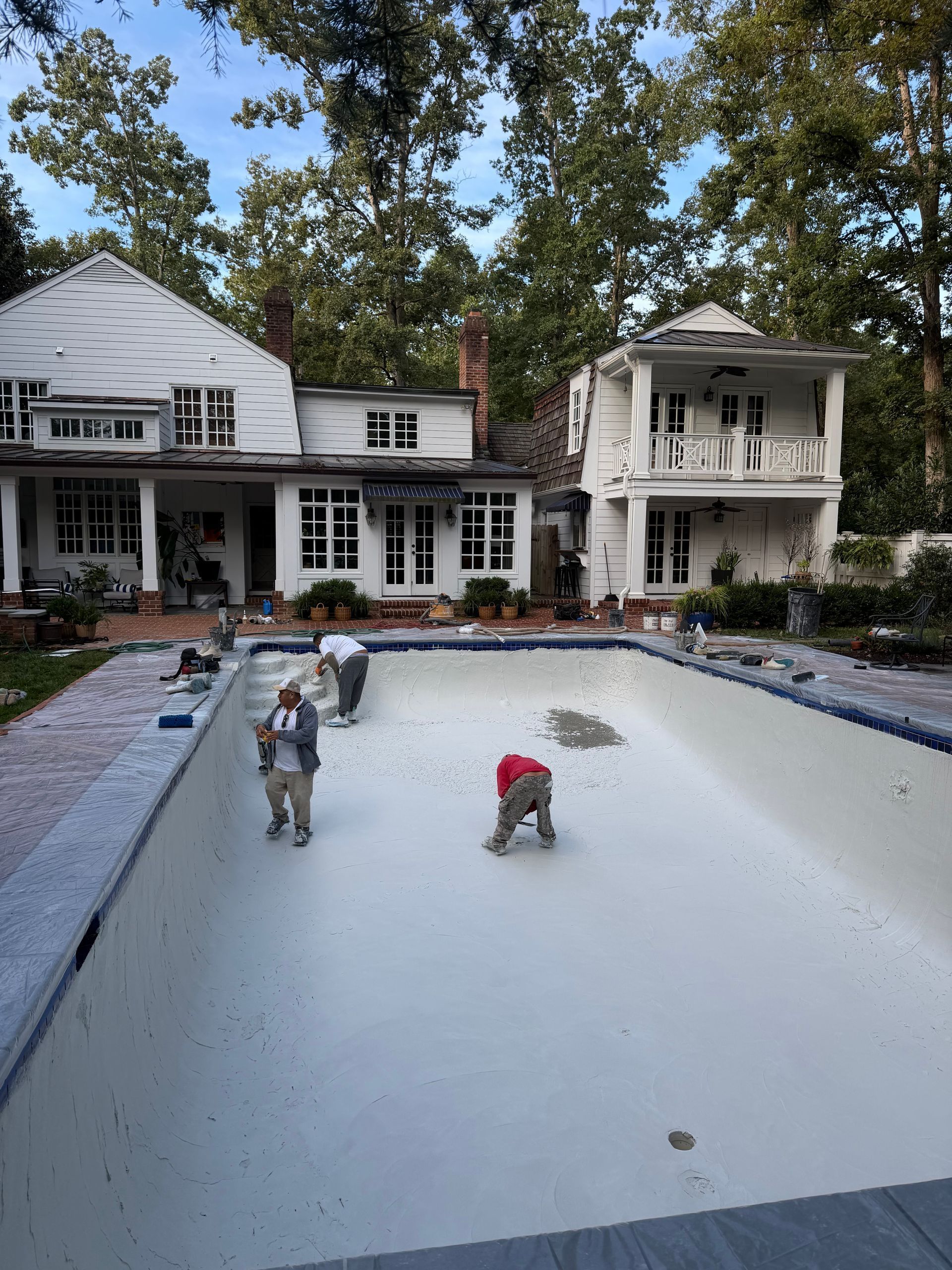 Workers resurfacing a backyard pool beside a white house and trees