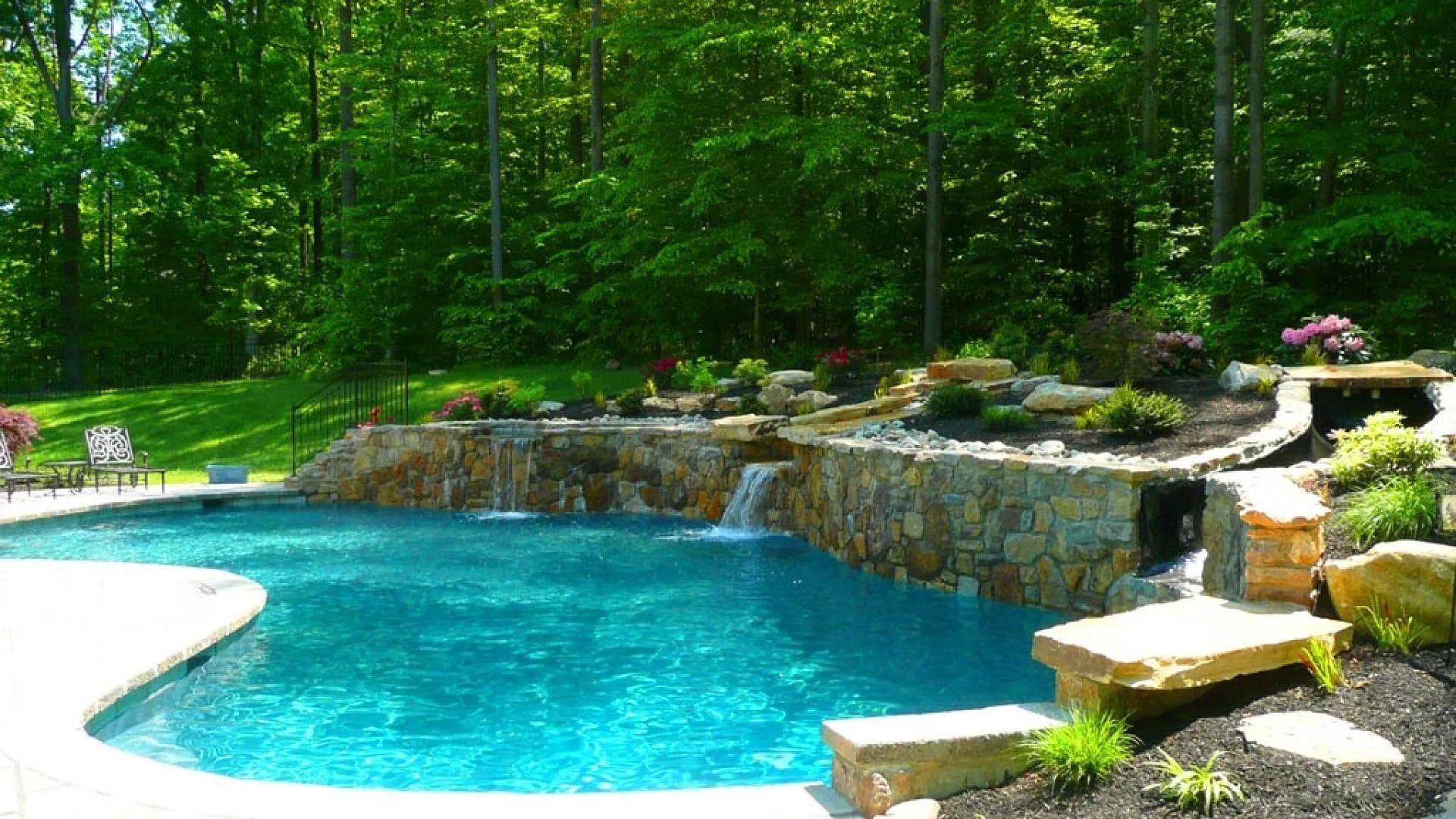 Swimming pool with a stone waterfall feature, surrounded by greenery and a forest backdrop.