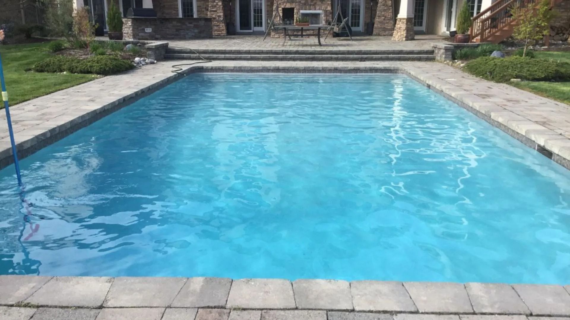 Rectangular blue swimming pool surrounded by stone pavers and green grass, with a building visible in the background.